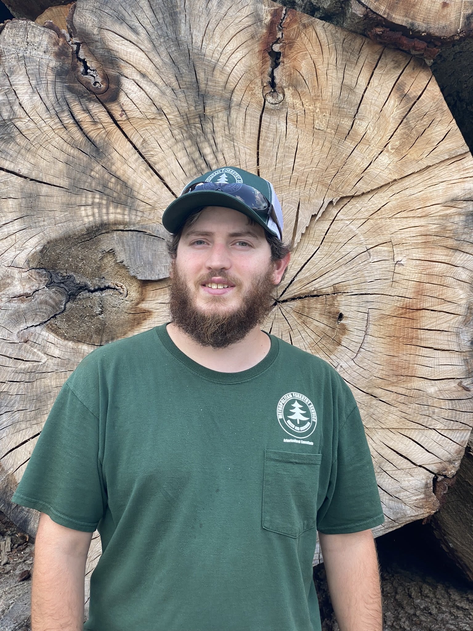 man with beard wearing a dark green hat and t-shirt standing in front of a tree trunk