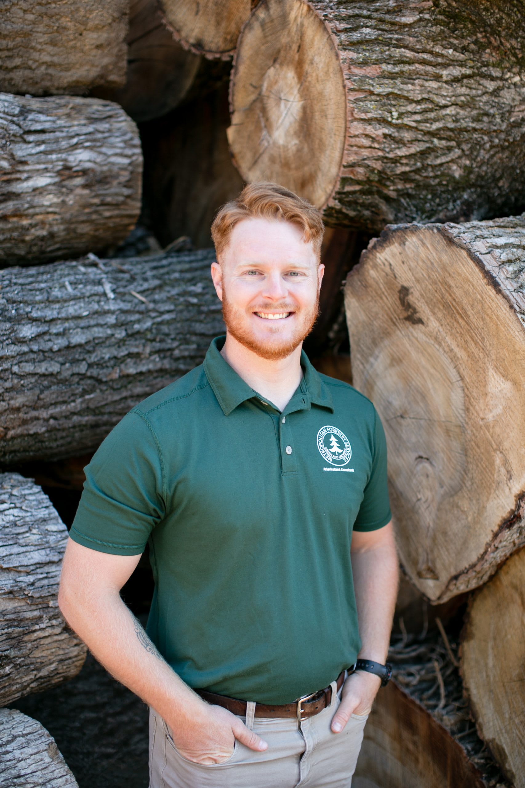 Man wearing a dark green shirt with his hands in his pockets standing in front of tree logs.