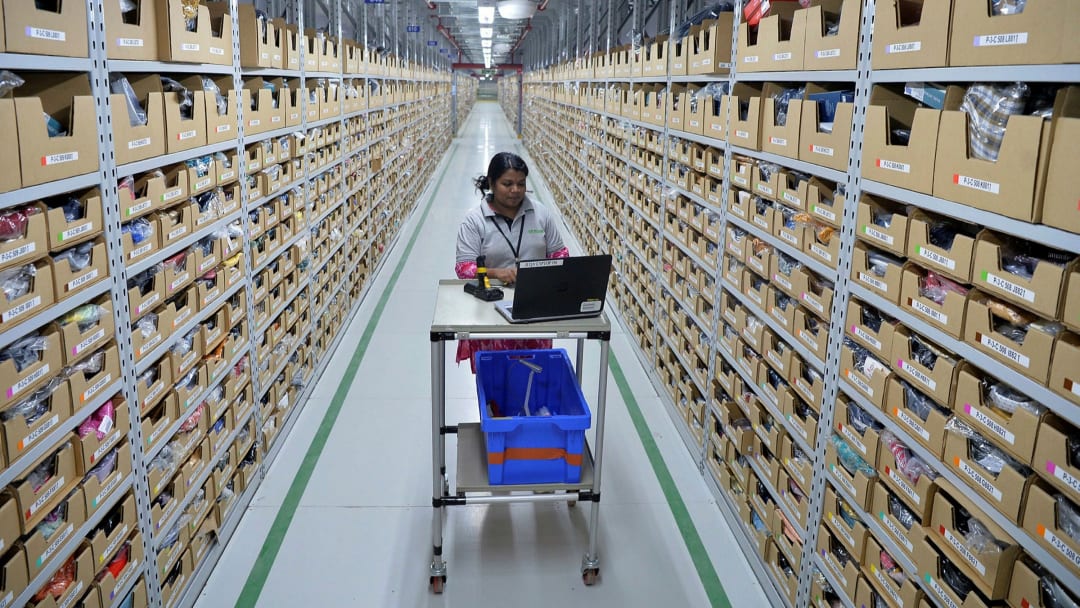 Warehouse worker with a laptop cart and blue tote picking orders in an aisle of labeled bin shelving