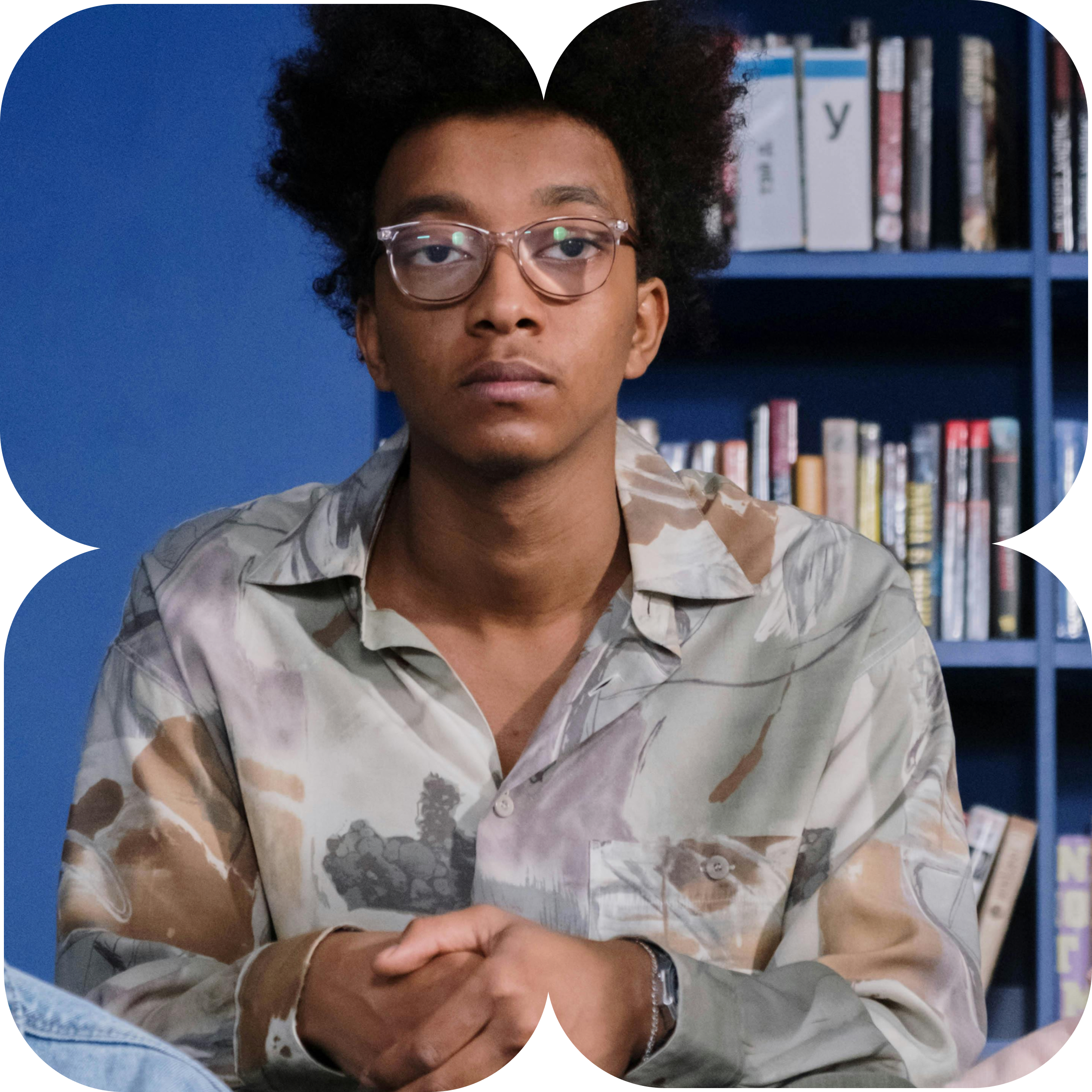 Un joven con gafas y cabello rizado sentado frente a una estantería con libros, con una expresión seria.