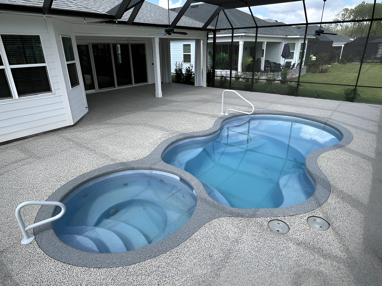 A textured pool deck featuring a hot tub and a clear blue pool under a screened enclosure.