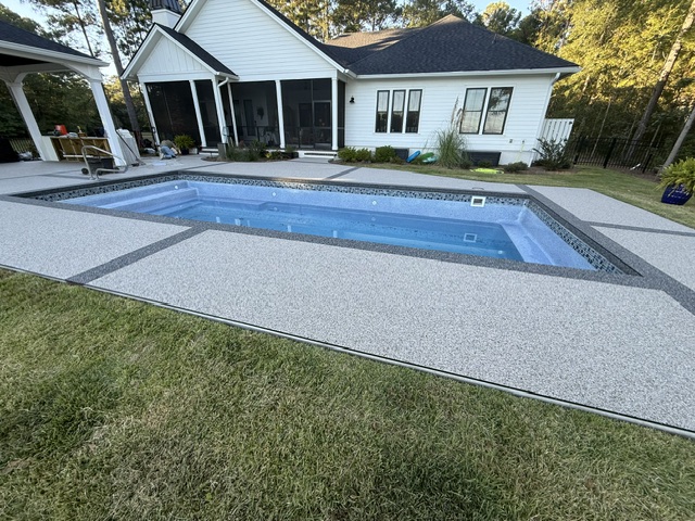 A resin-bound pool deck surrounding a clear blue pool with grassy areas and a house in the background.