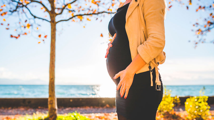 Pregnant woman holding belly wearing black and beige sweater in autumn outside near lake and tree