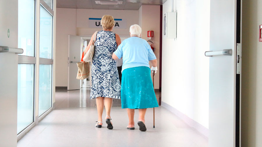 Elderly woman with swollen legs walking with cane in hospital and holding arm of another woman
