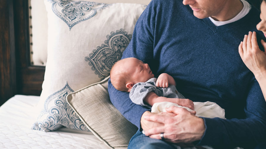 Father holding baby in arms while sitting on couch and mother is smiling