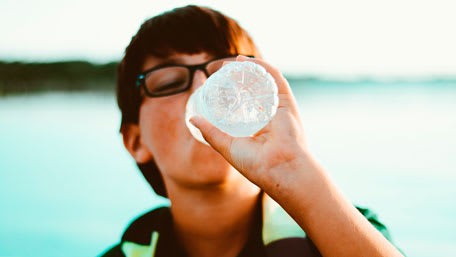 Boy with glasses drinking cold water from bottle with blurry ocean in background