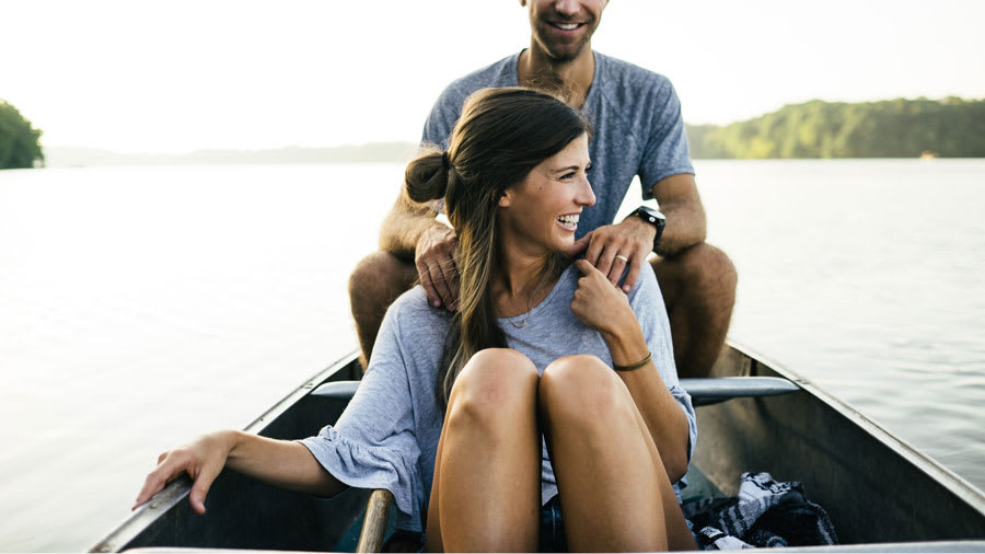 Brunette with clear face sitting on a canoe on the water with a man sitting behind her holding her shoulders