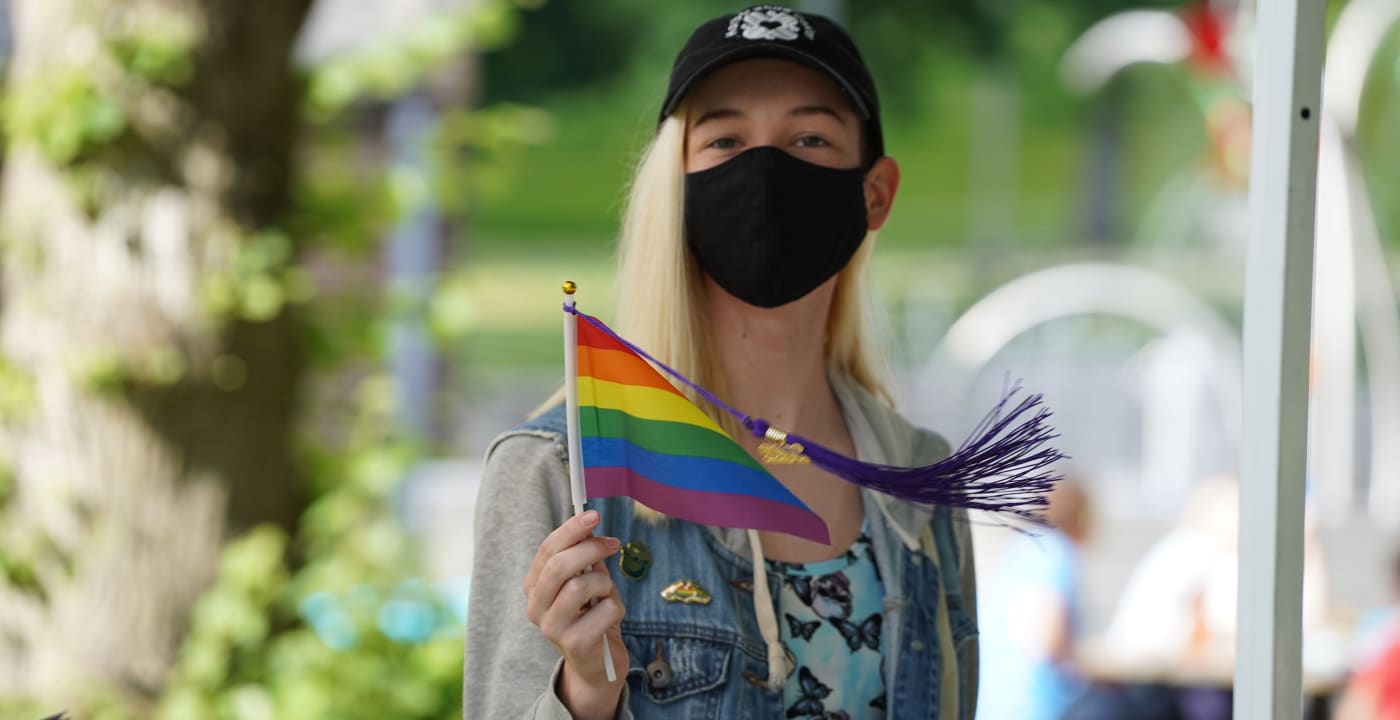 Person in a black ball cap and black face mask holds a rainbow Pride flag