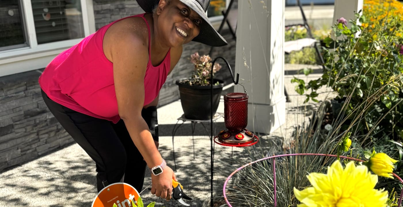 Woman in wide-brimmed hat holds plant clippers above a plant bed while smiling at the camera