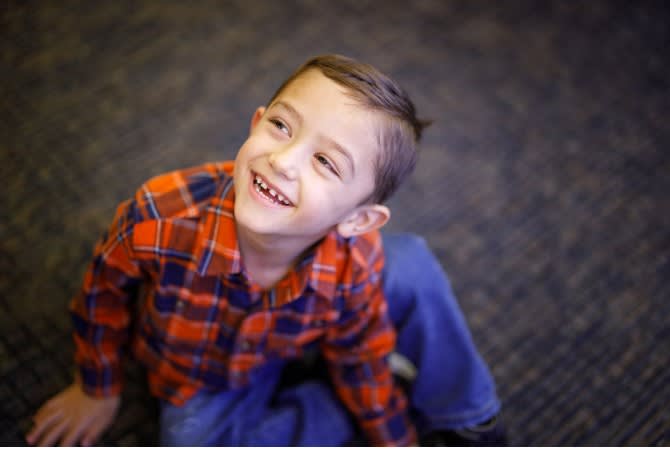 Young boy in a red-and-blue shirt sitting on the floor, looking up and smiling