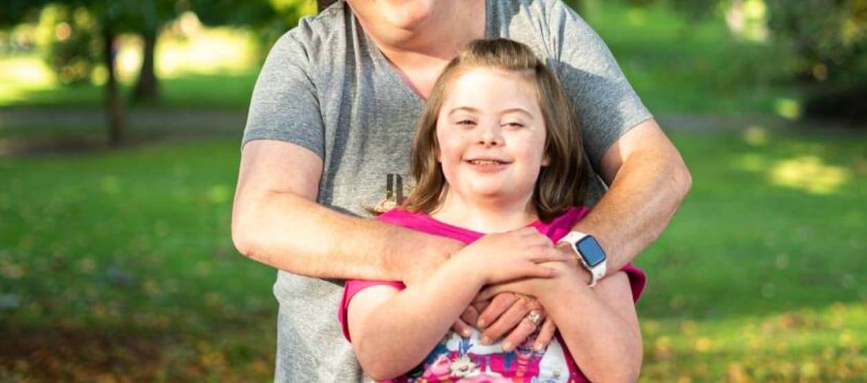 Pediatric cancer survivor Alexis smiles in a pink shirt and holds her mother’s hands, which are wrapped around her shoulders as she hugs Alexis from behind