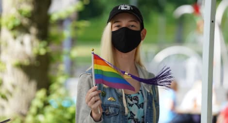 Person in a black ball cap and black face mask holds a rainbow Pride flag