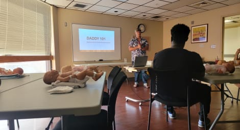 Man stands at front of classroom, “Daddy 101” presentation displayed on screen behind him; audience sits at tables with baby dolls