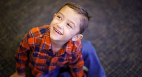 Young boy in a red-and-blue shirt sitting on the floor, looking up and smiling