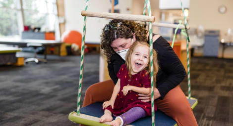 Smiling toddler wearing a red dress and purple orthotics on her left leg sits on a large swing with female physical therapist wearing a black top and orange pants