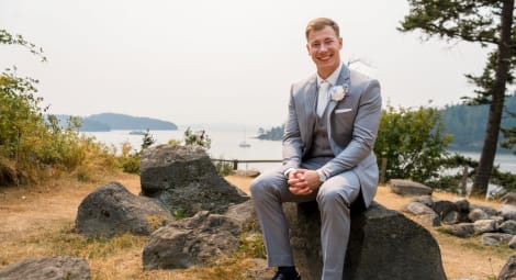 Young man wearing a gray suit sitting on a rock by the waterfront