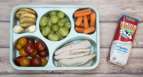 A bento box featuring rolled-up turkey slices, fruits, vegetables and a carton of milk.