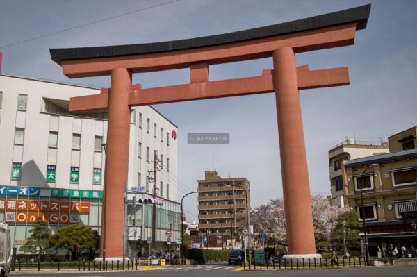 Enormous vermilion torii gate at a Nagoya street intersection framed by cherry blossoms and urban buildings