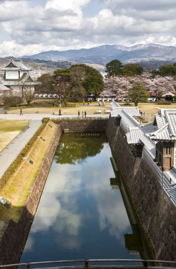 Kanazawa Castle's inner moat mirrors white-walled towers and turrets under a spring sky with cherry blossoms