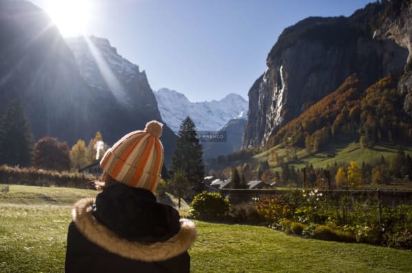 Figure in a winter jacket sits on a hillside looking over Lauterbrunnen Valley with sun rays and snow-capped Alps beyond