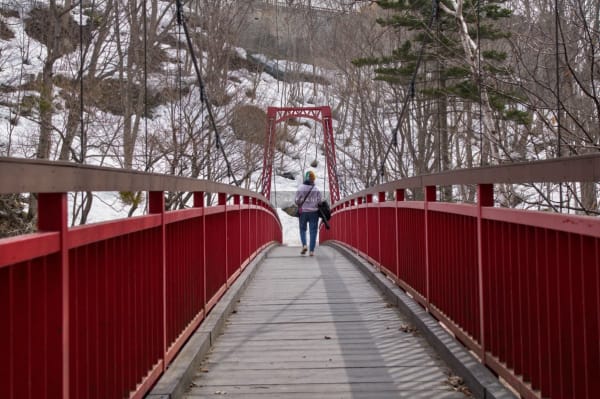 A lone figure crosses a red suspension bridge in a snow-covered winter landscape in Sapporo