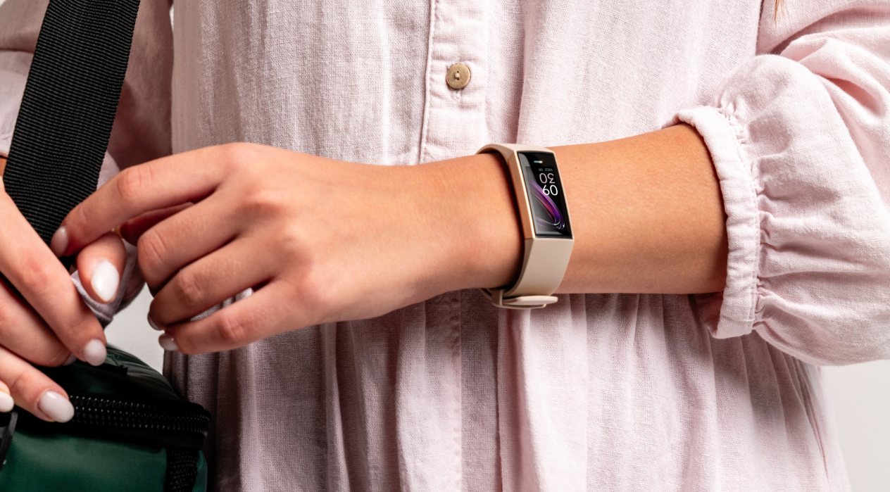 A person wearing a light, buttoned dress holds a green bag with black straps. They are wearing a smartwatch, and the background is plain.