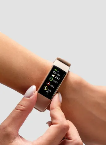 A smartwatch displays fitness metrics like steps and heart rate on the screen. A person is adjusting it on their wrist in a neutral, gray-background setting.