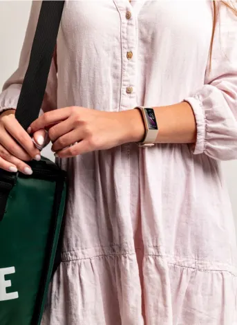 A person wearing a light, buttoned dress holds a green bag with black straps. They are wearing a smartwatch, and the background is plain.