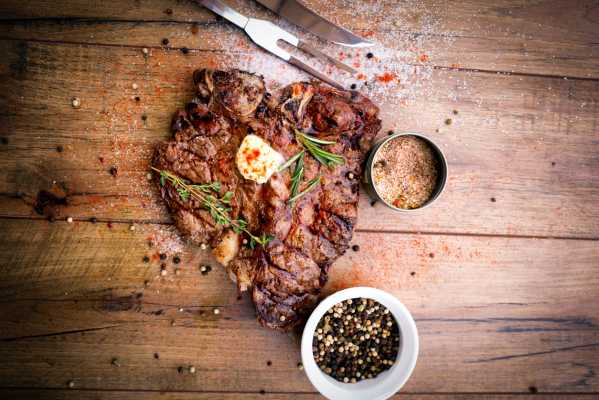 Perfectly grilled T-bone steak on a rustic cutting board with shallow depth of field and fresh thyme and rosemary and a pat of melted butter on top.