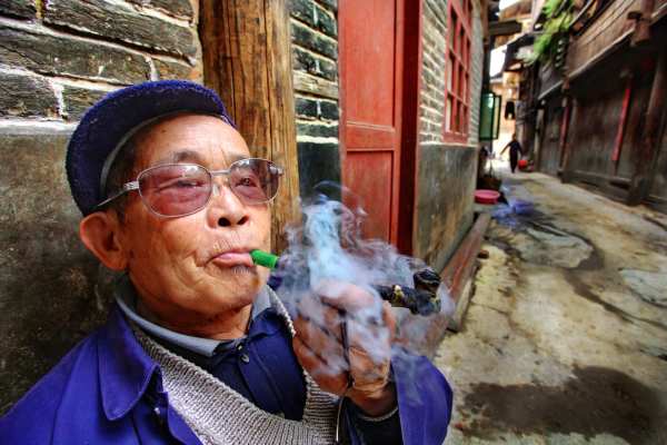 "Zhaoxing village, Guizhou province, China - April 9, 2010:  An elderly Chinese farmer wearing glasses, smoking a pipe outside his home April 9, 2010. Zhaoxing Dong villages, Liping County. Chinese peasant produces tobacco smoke."