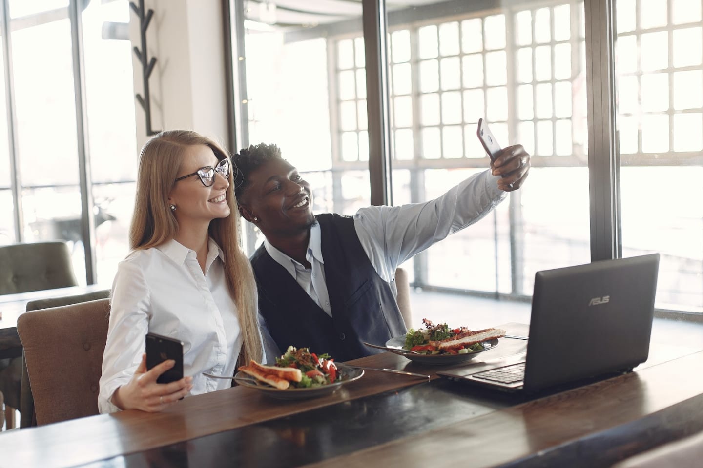 Two professionally dressed people sit next to eachother, taking a selfie.