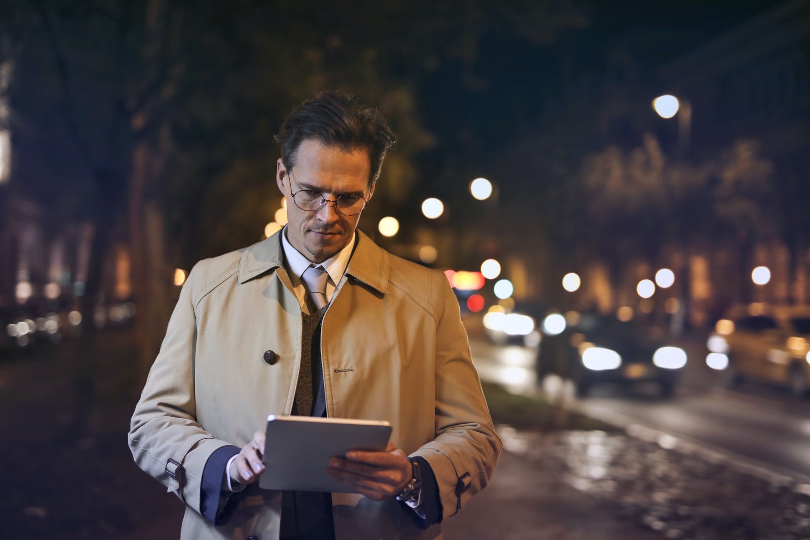 A smartly dressed man uses a tablet computer while walking down a city street at night.