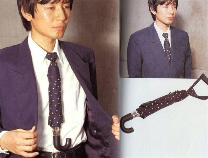 A man in a business suit wears a tie that appears to also be an umbrella. He opens his jacket to reveal an umbrella handle projecting from the end of his tie.