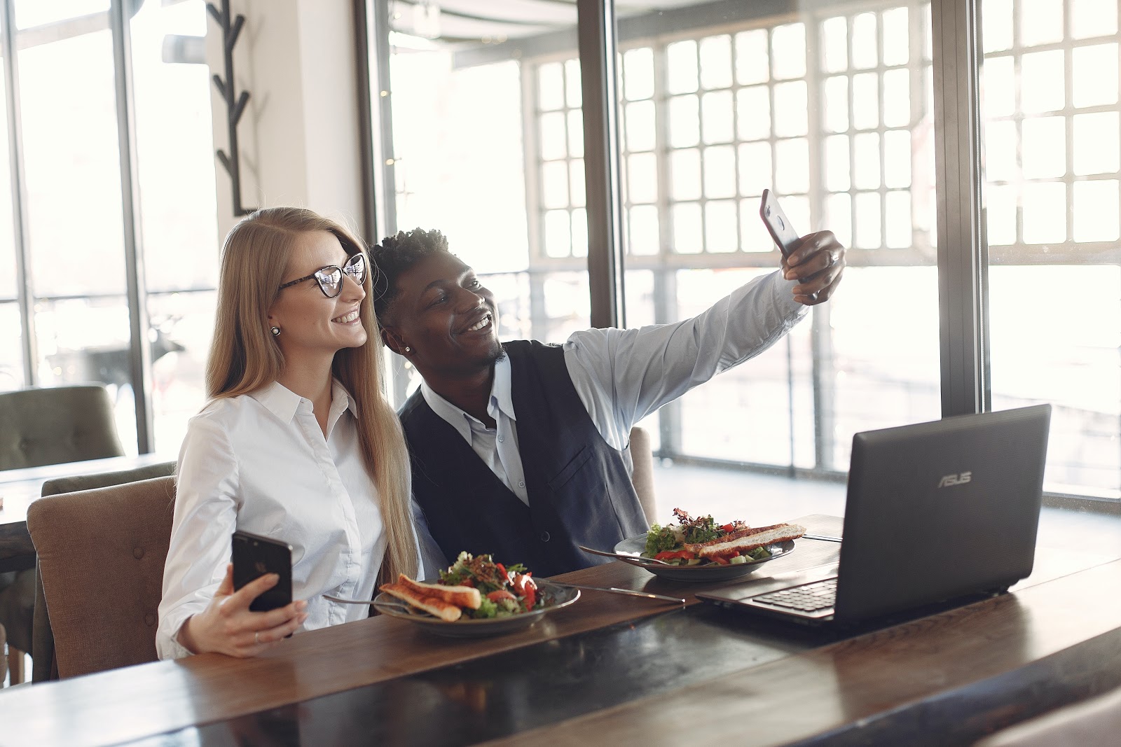 Two professionally dressed people sit next to eachother, taking a selfie.