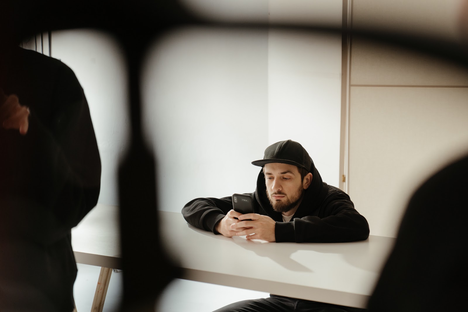A man in a black hoodie and baseball cap sits at a desk and gazes into the phone he holds in both hands.