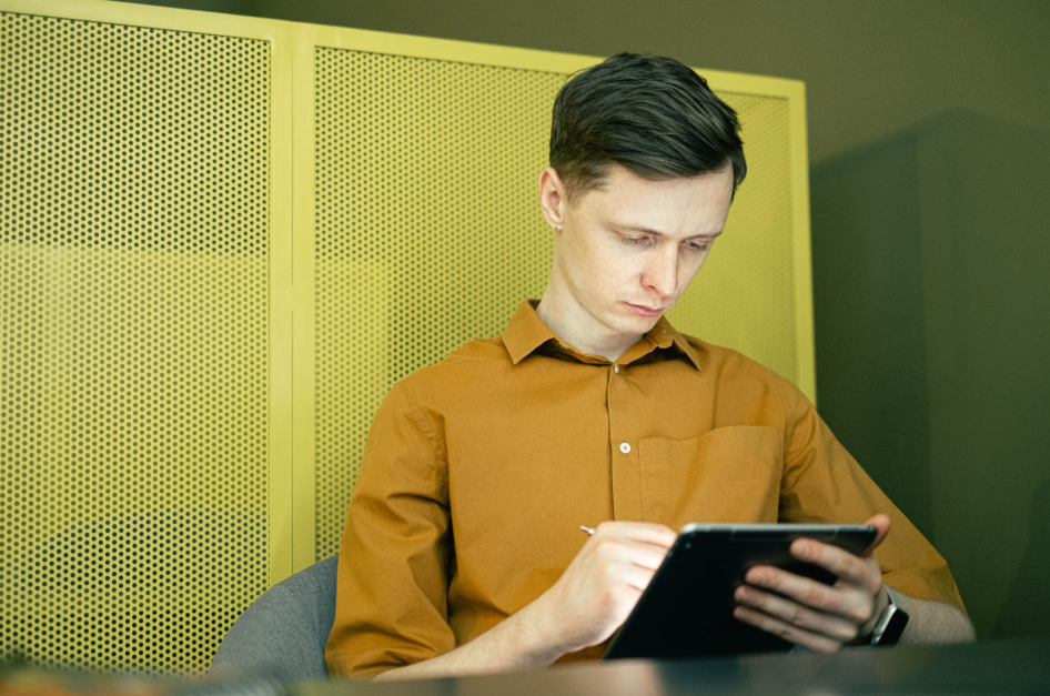 A man in a mustard yellow shirt reads from a tablet computing device.
