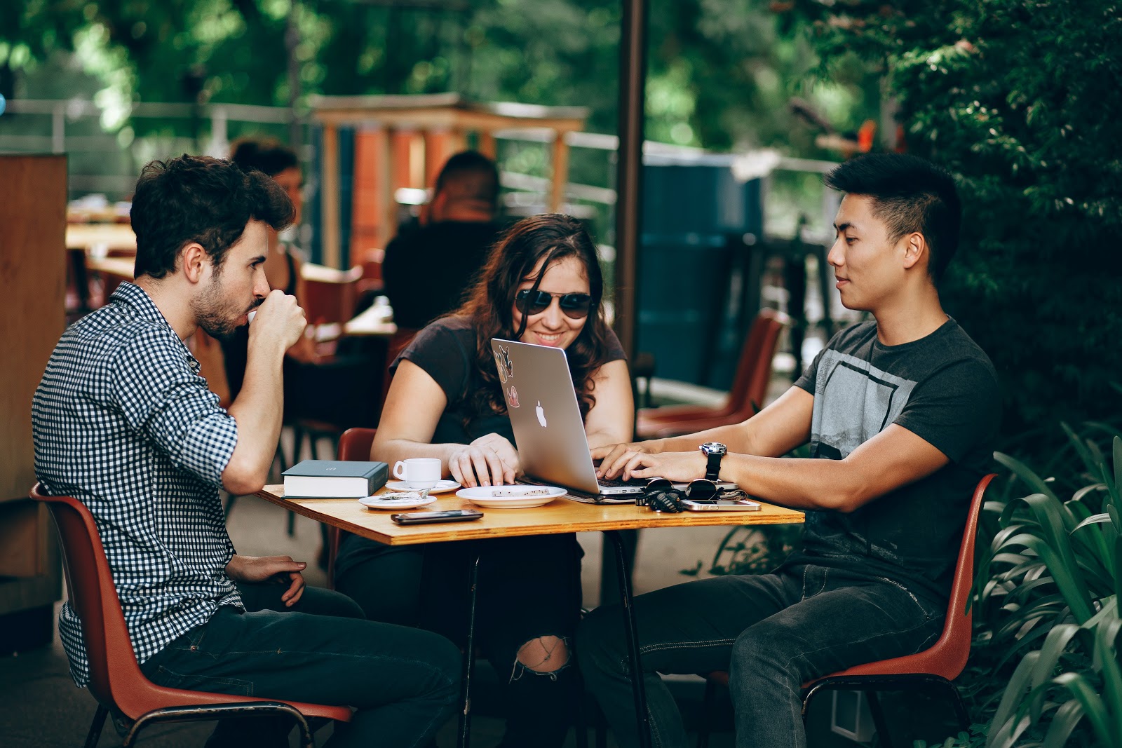 A group of young, casually dressed people work together at an outdoor cafe table, laptops open and smiling in the sun.
