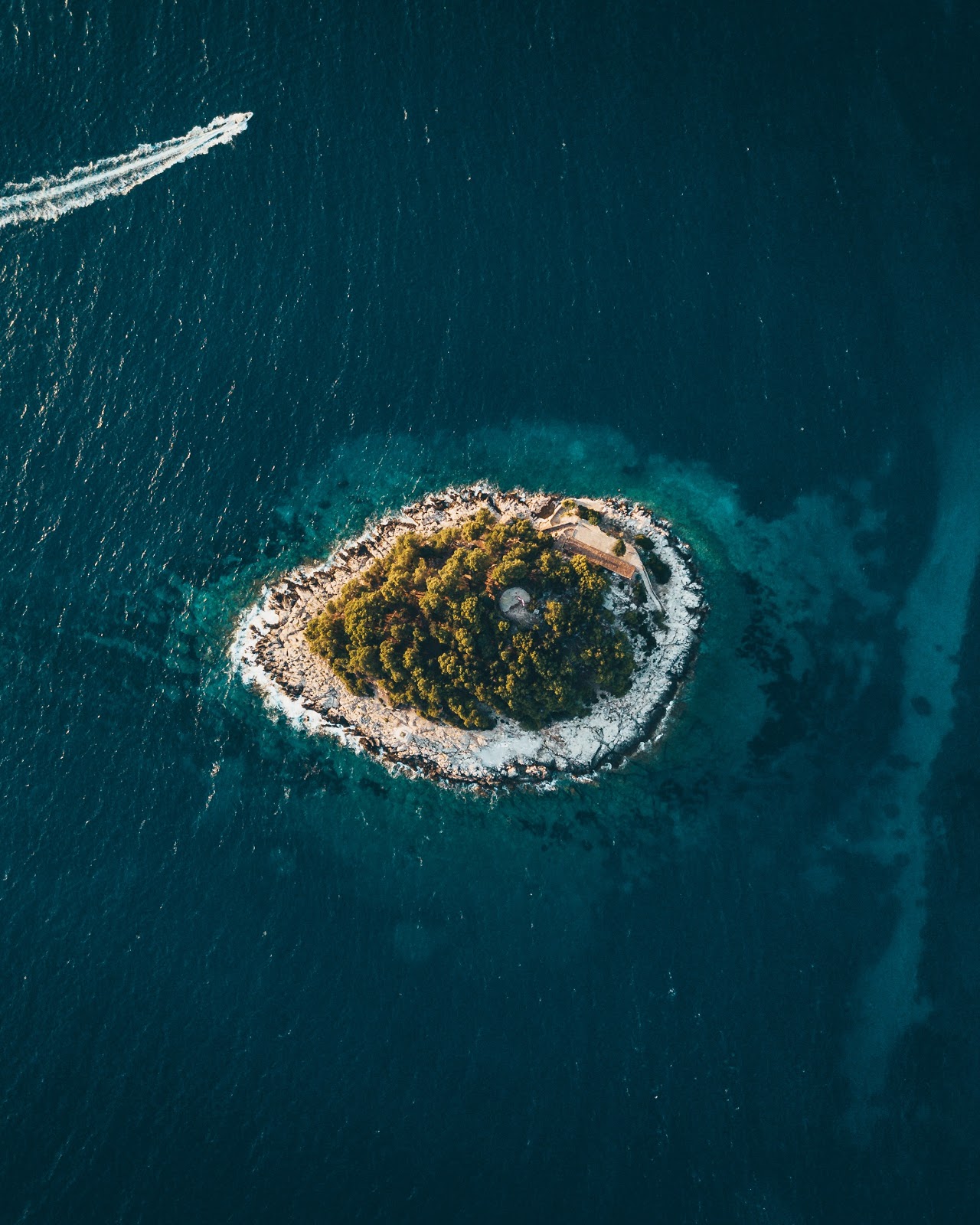 A beautiful island rich with foliage and surrounded by a white shingle sits in the midst of a sparkling azure sea. Toward the top of the frame, a speedboat is passing.