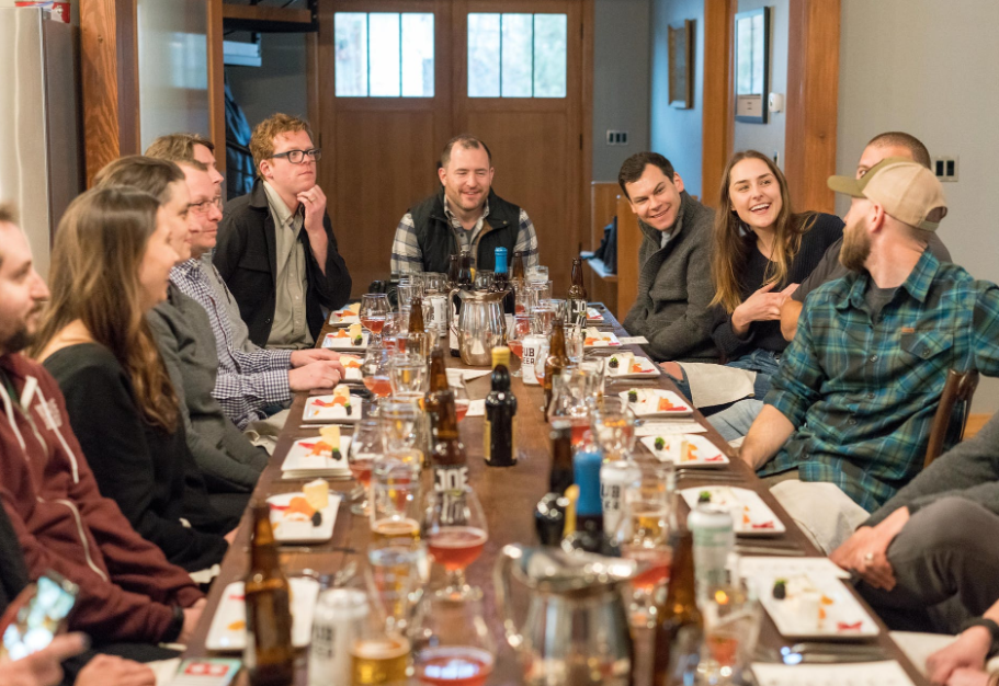 A group of perhaps a dozen people in smart-to-smart casual attire sit around a table laden with food and drink, enjoying a discussion.