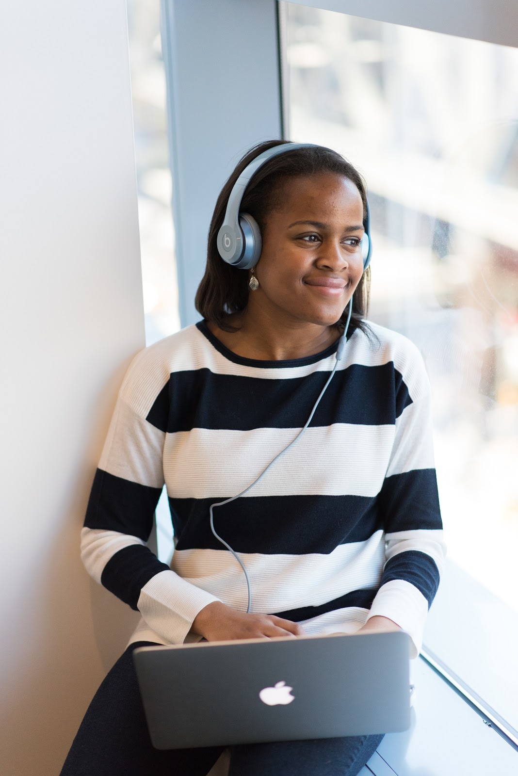 A young woman wearing large headphones listens to music on her laptop while happily gazing out of the window.