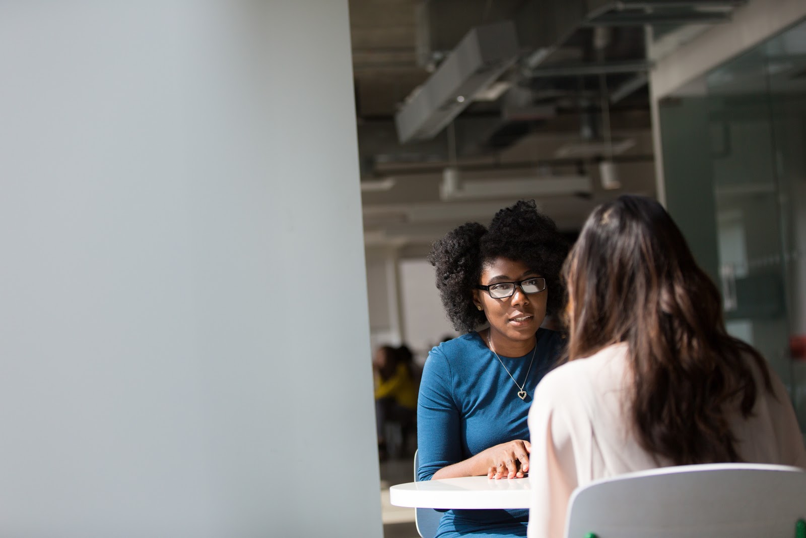 Two women sit in an interview like scenario at a table. One has her back toward us.