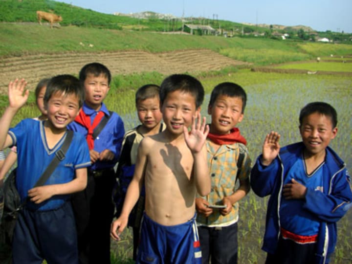 Seven children stand in a field waving at the camera. Behind them, there are rolling hills with crop lines and a cow.