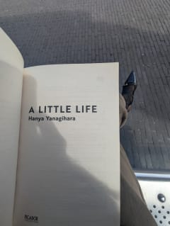 Olu waiting at the bus stop, holding a copy of A little life