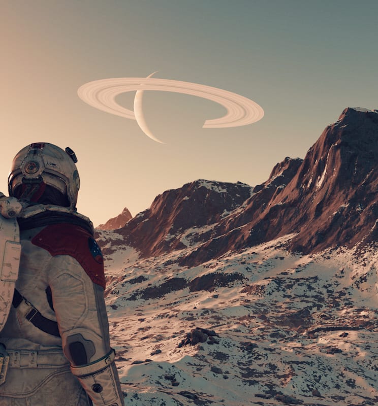 A space explorer looks out over a snowy mountain range on a strange planet. A ringed planet hangs on the horizon. 