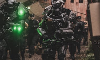 Armored Enclave soldiers with glowing green lights advance through a forest path toward a wooden outpost.