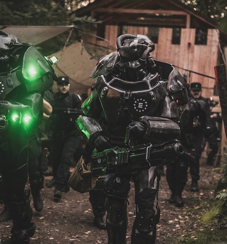 Armored Enclave soldiers with glowing green lights advance through a forest path toward a wooden outpost.