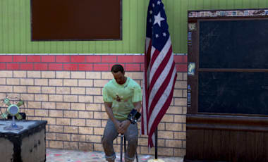 A C.A.M.P. in Fallout 76 is designed to look like a classroom. A character in a green t-shirt and jeans sits on a stool. Behind them in a chalk board, a flag, a desk, and a skeleton