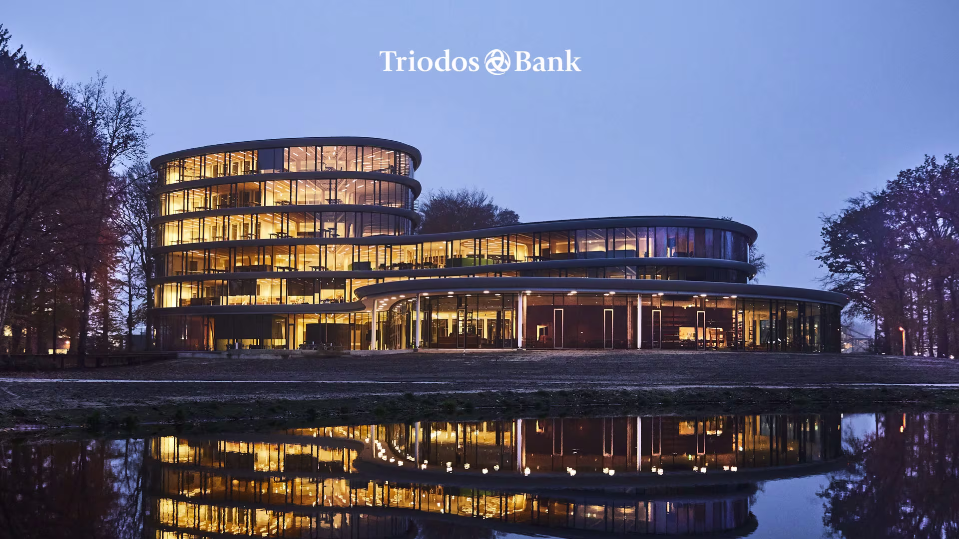 Modern kantoorgebouw van Triodos Bank bij schemering, verlicht met warm licht, weerspiegeld in water met bomen eromheen.