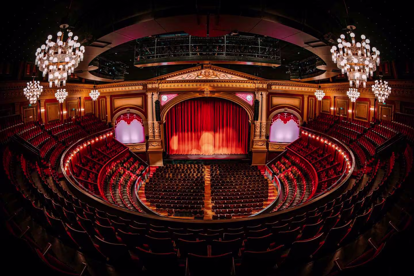 Luxueus theaterinterieur met rode gordijnen, kristallen kroonluchters en rijen rode stoelen in een ovale zaal.