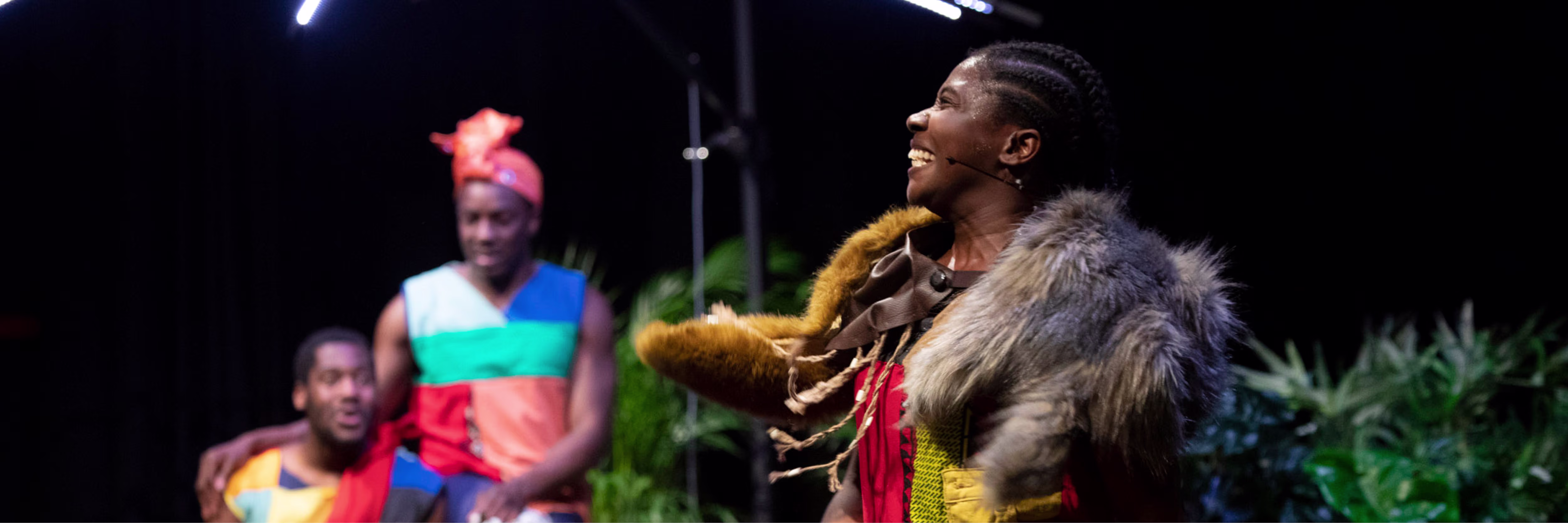 Een acteur in traditionele kleding met bontjas speelt op het toneel, met andere performers op de achtergrond in kleurrijke kostuums.